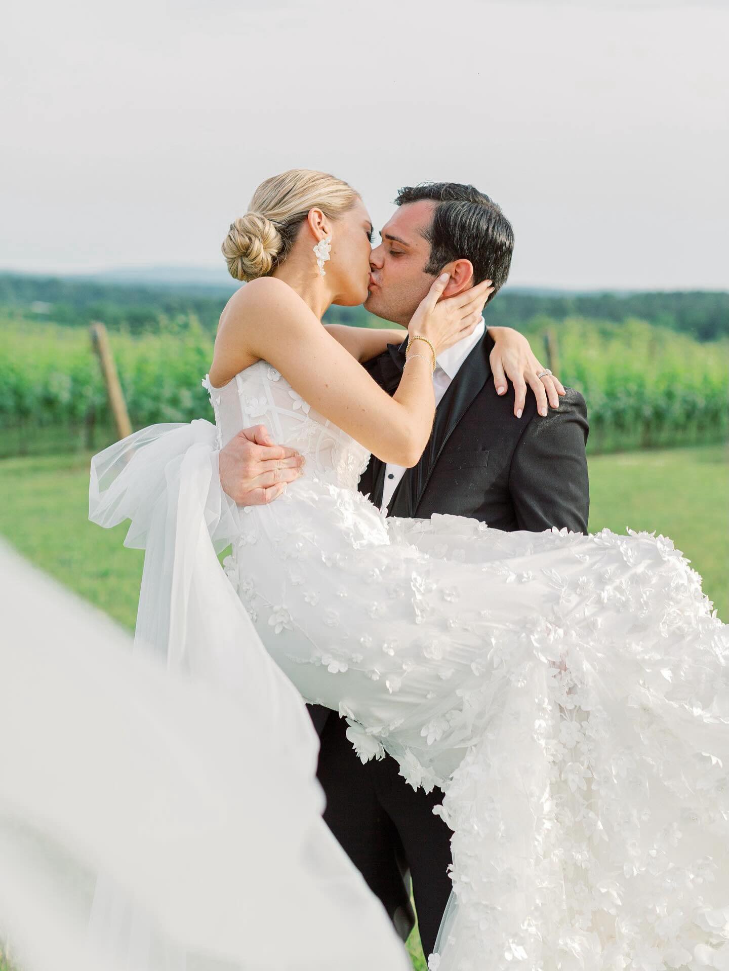 Groom carrying bride in floral-embellished wedding gown while kissing at vineyard with green hills beyond