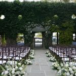 Wedding ceremony site with hedge wall, pavilion entrance, and formal seating arranged around white floral displays