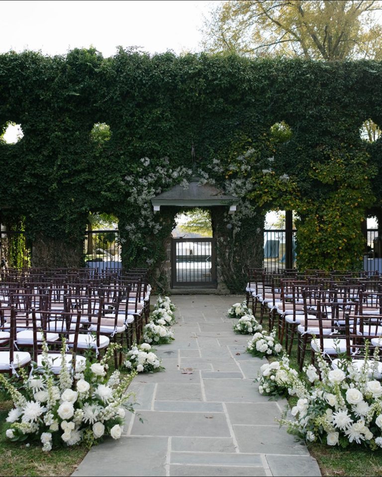 Wedding ceremony site with hedge wall, pavilion entrance, and formal seating arranged around white floral displays
