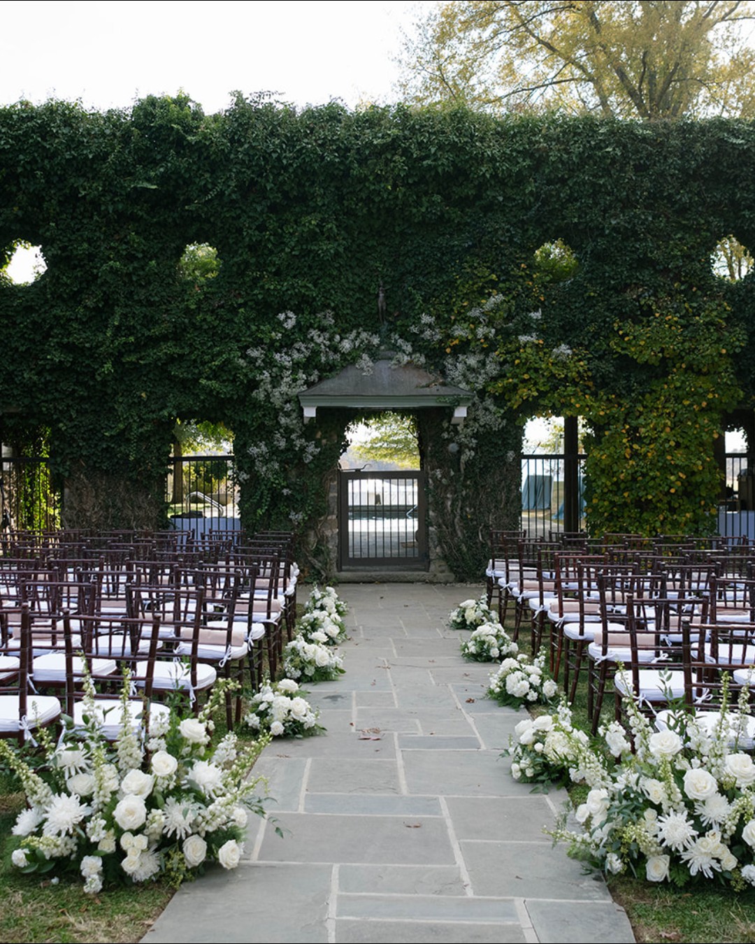 Wedding ceremony site with hedge wall, pavilion entrance, and formal seating arranged around white floral displays
