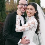 Newlywed couple embracing outdoors, groom in black tuxedo and bride in lace gown with veil