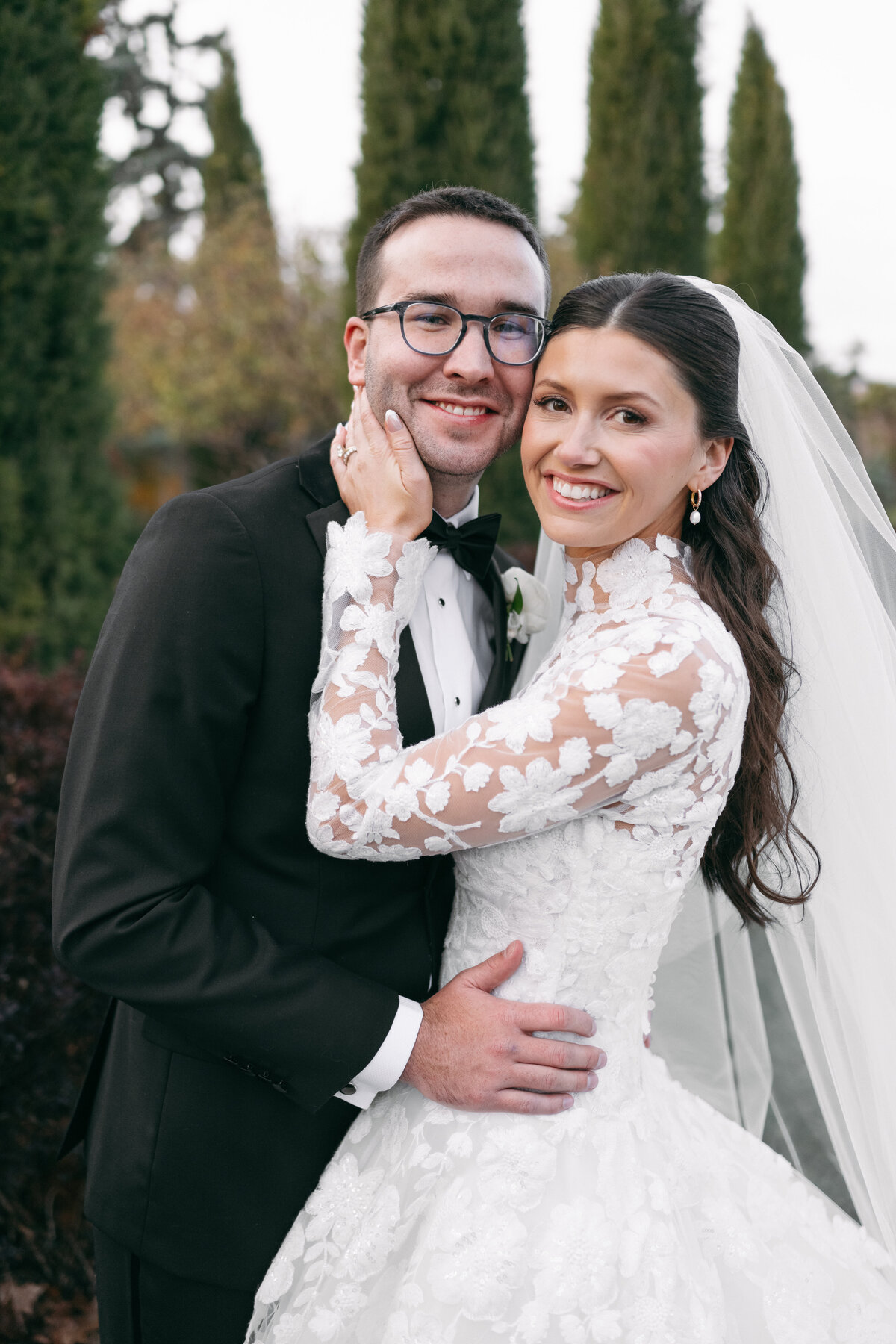 Newlywed couple embracing outdoors, groom in black tuxedo and bride in lace gown with veil