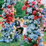 Wedding couple framed by colorful floral ceremony arch featuring blue hydrangeas, pink and coral blooms