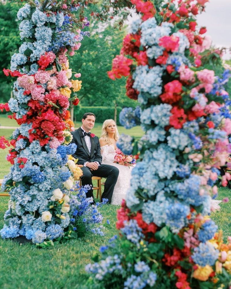 Wedding couple framed by colorful floral ceremony arch featuring blue hydrangeas, pink and coral blooms