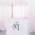 White mobile bar cart with custom monogram under fringed umbrella at outdoor wedding reception