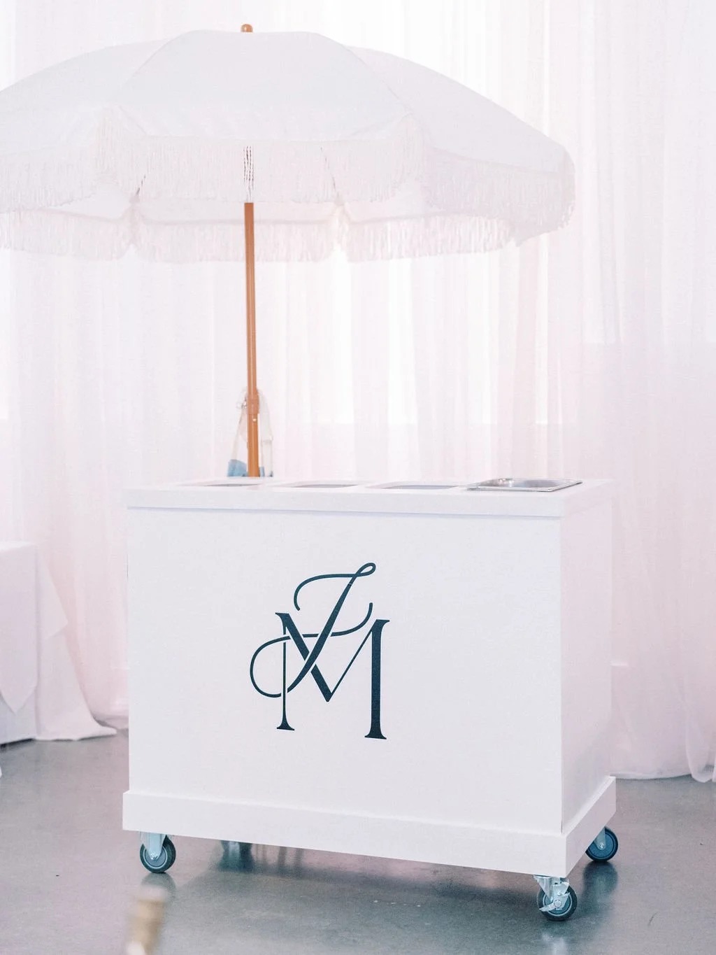 White mobile bar cart with custom monogram under fringed umbrella at outdoor wedding reception