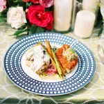 Plated dinner featuring chicken, roasted vegetables, and salmon on decorative blue plate with floral centerpiece and candles