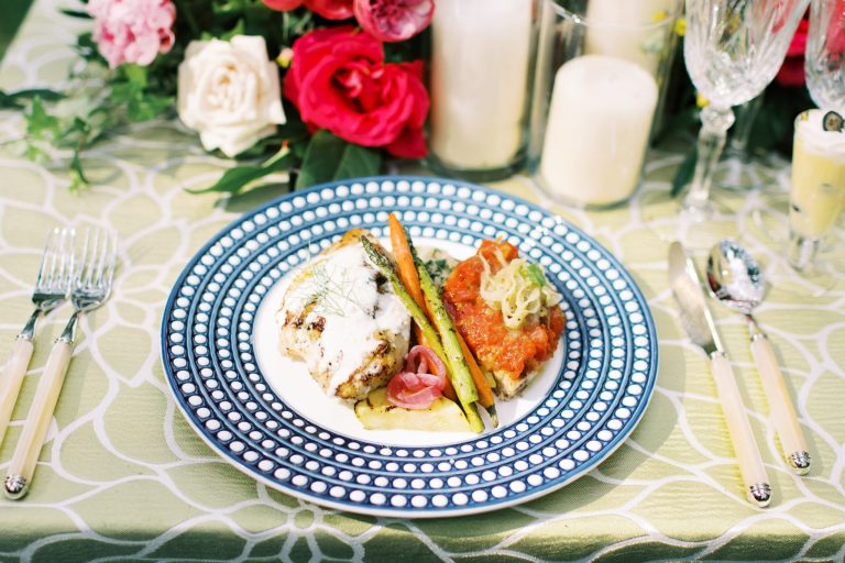 Plated dinner featuring chicken, roasted vegetables, and salmon on decorative blue plate with floral centerpiece and candles
