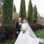 Newlyweds posing together outdoors with bride in lace gown with cathedral veil and groom in black tuxedo