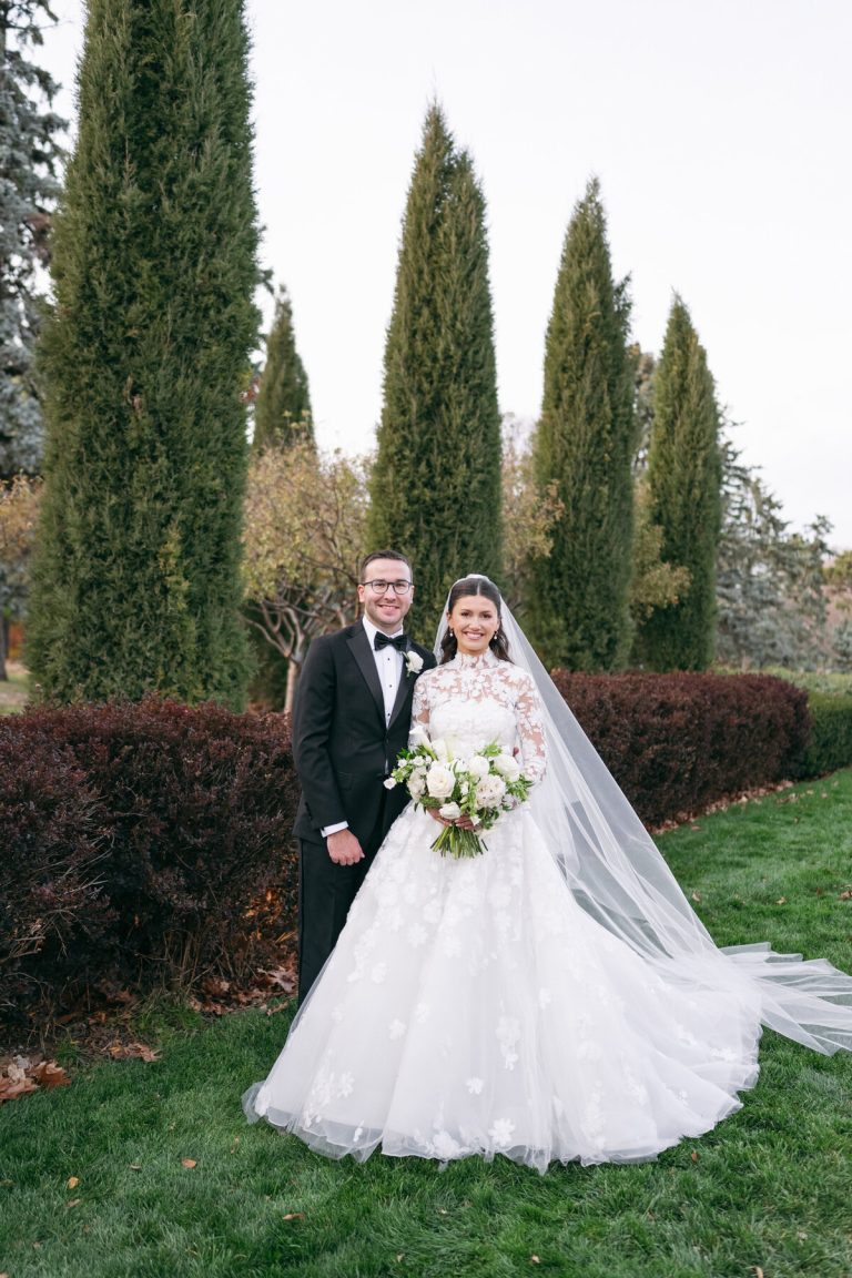 Newlyweds posing together outdoors with bride in lace gown with cathedral veil and groom in black tuxedo