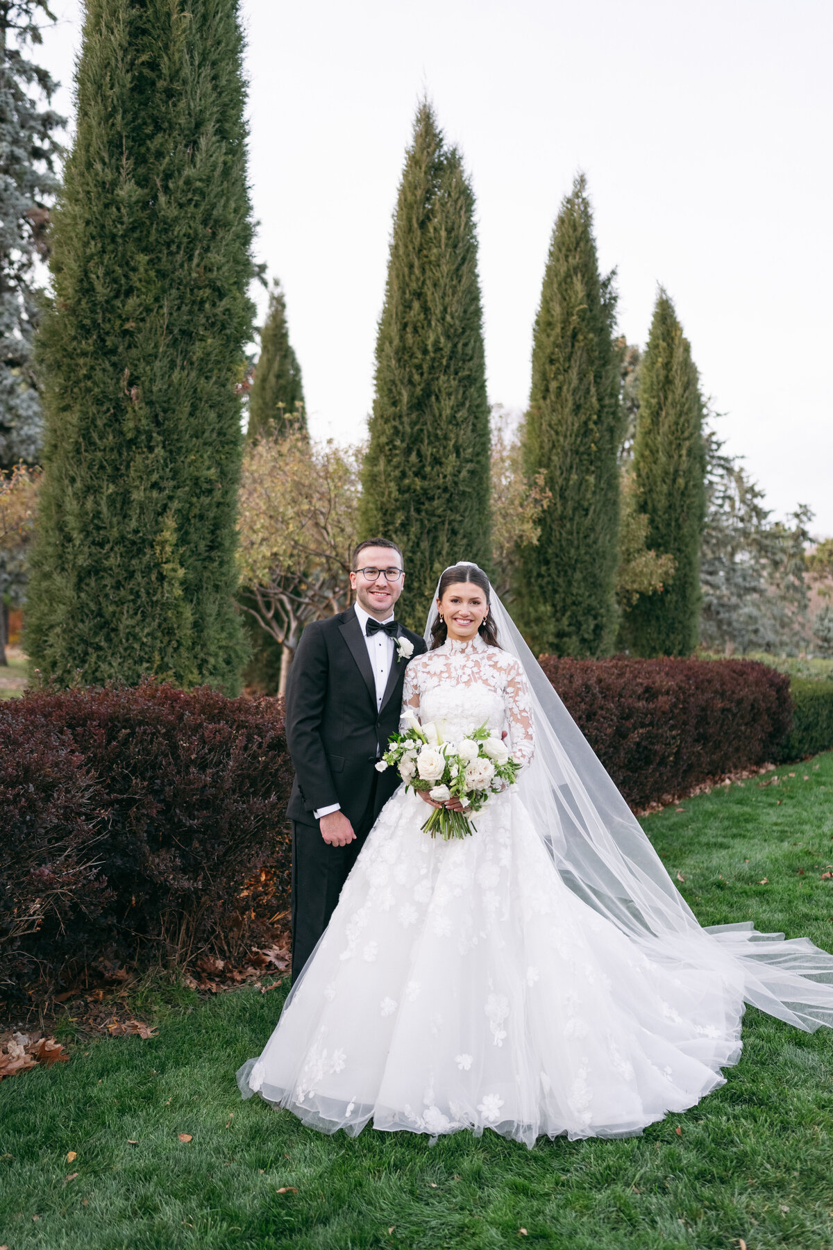 Newlyweds posing together outdoors with bride in lace gown with cathedral veil and groom in black tuxedo