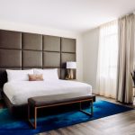 Contemporary hotel room with tufted headboard, white bedding, blue area rug, and gold-framed mirror
