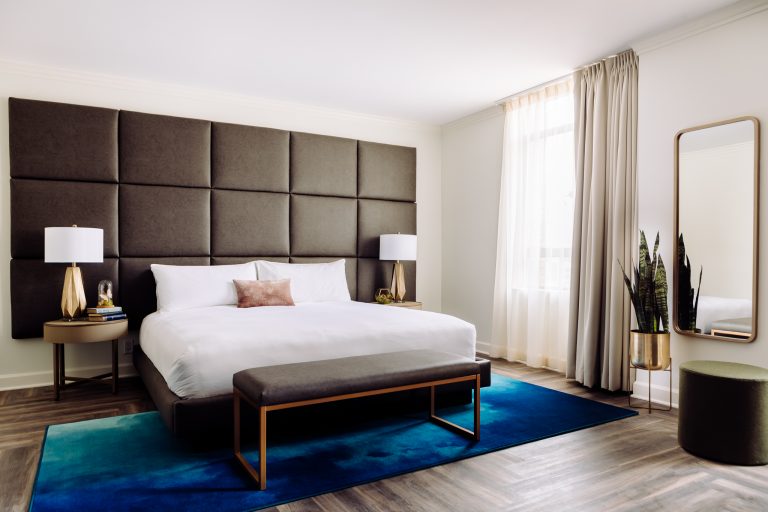 Contemporary hotel room with tufted headboard, white bedding, blue area rug, and gold-framed mirror