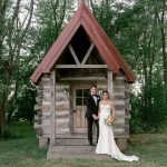 Newlyweds posing on wooden porch of rustic log cabin chapel with distinctive red A-frame roof and forest backdrop