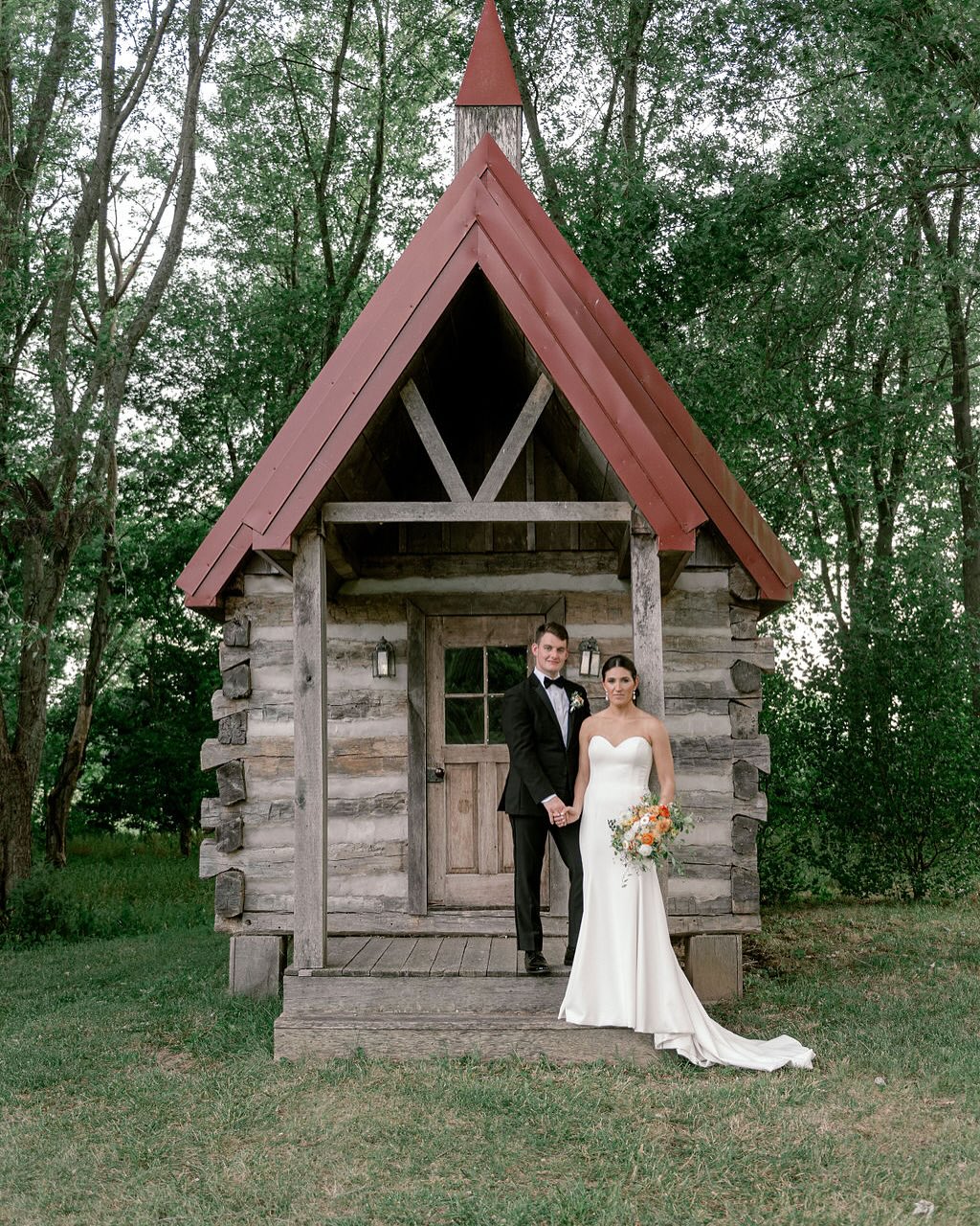 Newlyweds posing on wooden porch of rustic log cabin chapel with distinctive red A-frame roof and forest backdrop