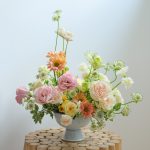 Pastel spring wedding centerpiece with roses, gerbera daisies, and delicate greenery in a blue ceramic compote vase