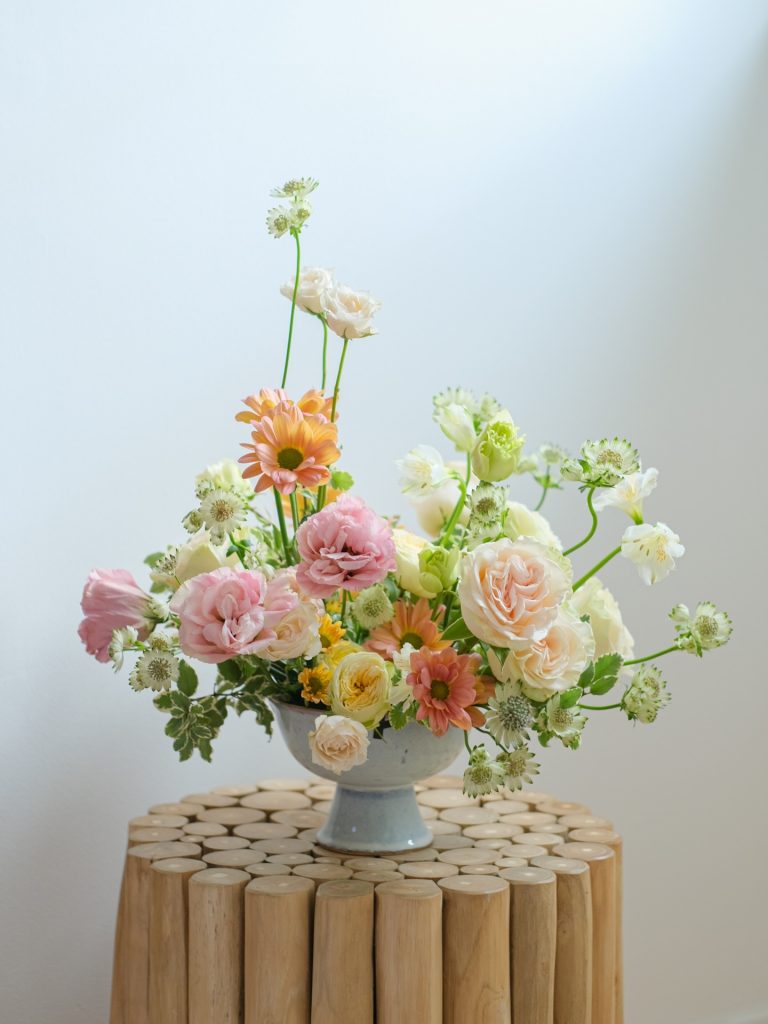 Pastel spring wedding centerpiece with roses, gerbera daisies, and delicate greenery in a blue ceramic compote vase