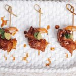 Three glazed chicken skewers garnished with cilantro and pineapple on a white woven plate
