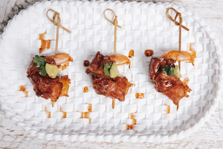 Three glazed chicken skewers garnished with cilantro and pineapple on a white woven plate