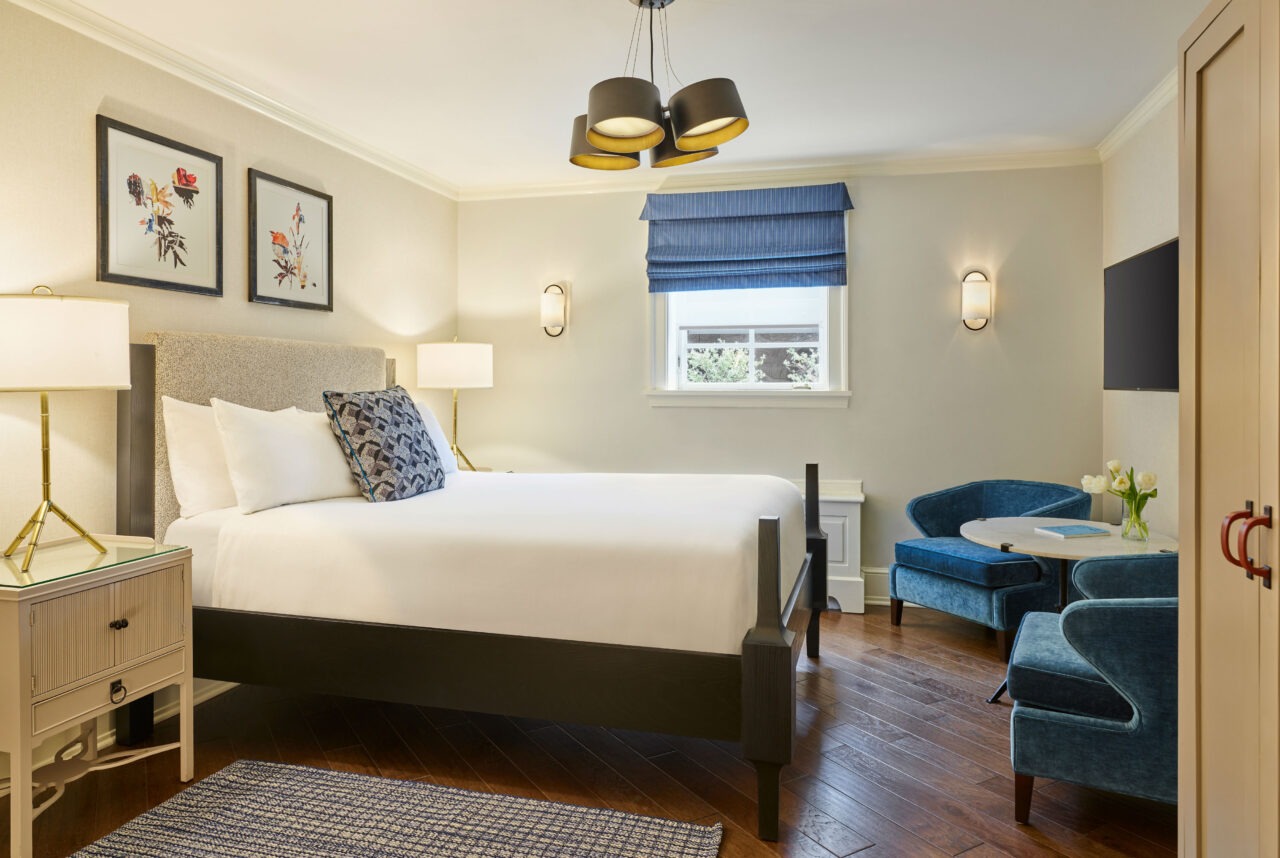 Bright hotel room with white bedding, blue velvet chairs, dark wood floors, and botanical wall art