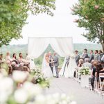 Jewish wedding ceremony under chuppah with officiant, bride and groom, and wedding party against scenic outdoor backdrop