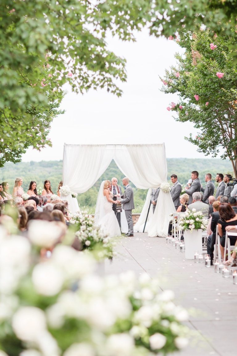 Jewish wedding ceremony under chuppah with officiant, bride and groom, and wedding party against scenic outdoor backdrop