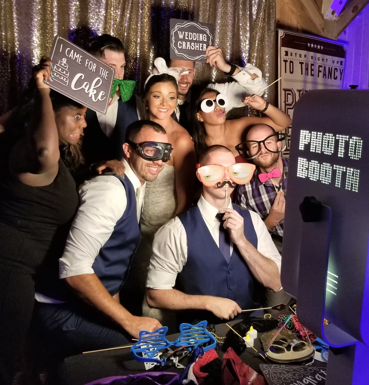 Wedding guests posing in photo booth with props and silly glasses against gold sequin backdrop