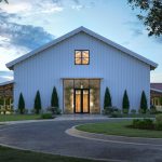 Modern white barn-style wedding venue with illuminated entrance, cypress trees, and circular driveway at dusk