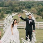 Newlyweds celebrating under white floral arch at outdoor countryside wedding ceremony
