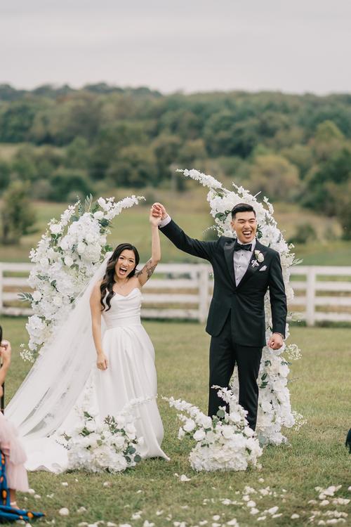 Newlyweds celebrating under white floral arch at outdoor countryside wedding ceremony