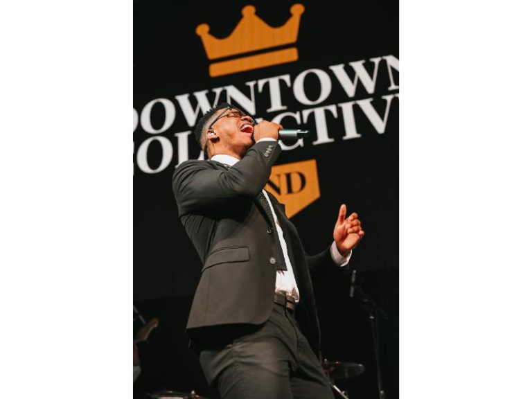 Male vocalist in suit singing passionately on stage with Downtown Collective branded backdrop