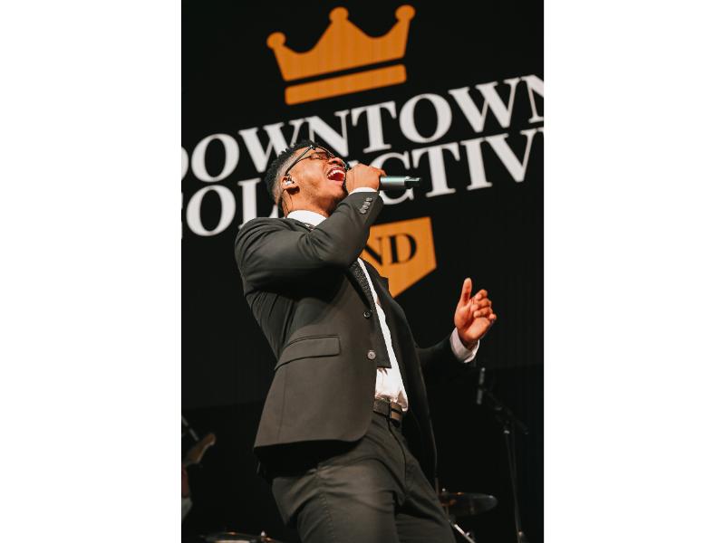 Male vocalist in suit singing passionately on stage with Downtown Collective branded backdrop