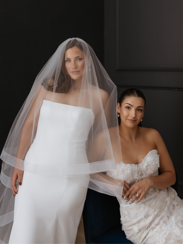 Two brides modeling wedding dresses, one in a minimalist strapless gown with veil, the other in a lace embellished dress
