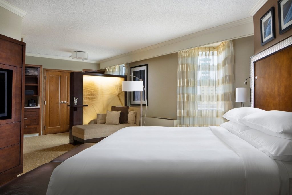 Spacious hotel suite with king bed, chaise lounge, wooden cabinetry, and neutral tones