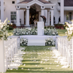 Outdoor wedding ceremony setup with white floral arrangements, petal-lined aisle, and colonial-style clubhouse