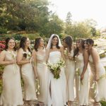 Bride in white strapless gown with bridesmaids in champagne dresses holding white floral bouquets in garden setting