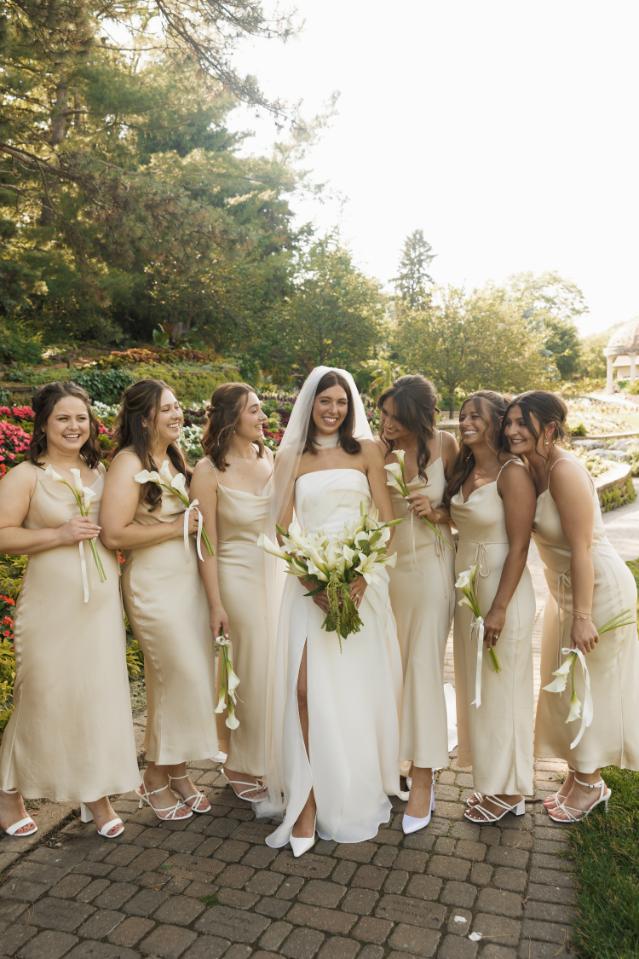 Bride in white strapless gown with bridesmaids in champagne dresses holding white floral bouquets in garden setting