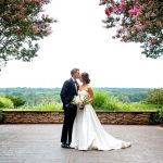 Bride in strapless white gown and groom in navy suit kiss beneath pink flowering trees overlooking valley