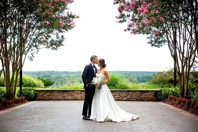Bride in strapless white gown and groom in navy suit kiss beneath pink flowering trees overlooking valley