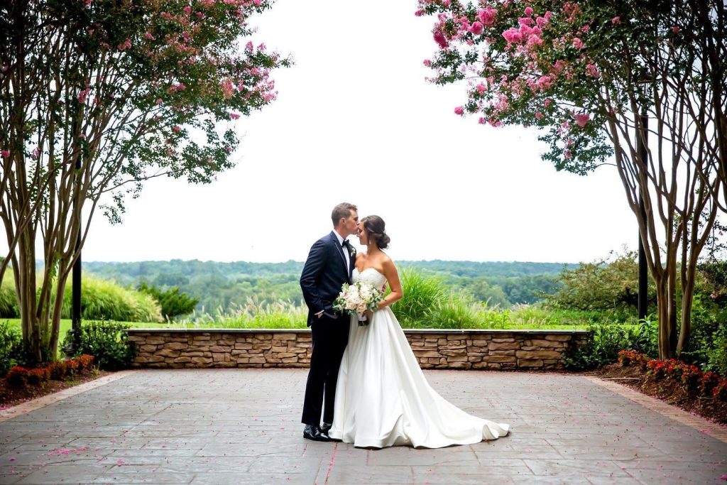 Bride and groom kissing on stone patio with flowering crepe myrtle trees and rolling hills in background