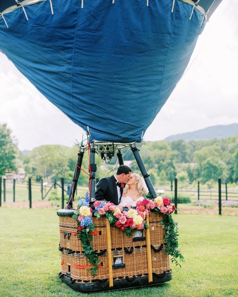 Bride and groom kissing in hot air balloon basket decorated with vibrant pink, blue, and yellow floral garlands
