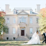 Newlyweds walking in front of white colonial mansion in autumn