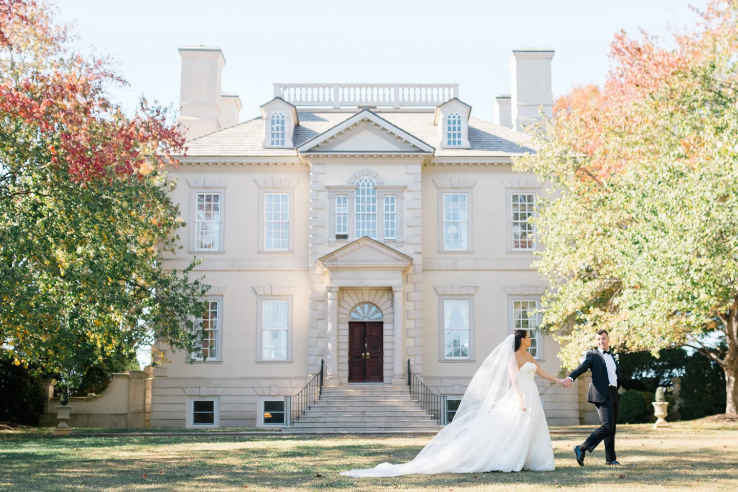 Newlyweds walking in front of white colonial mansion in autumn