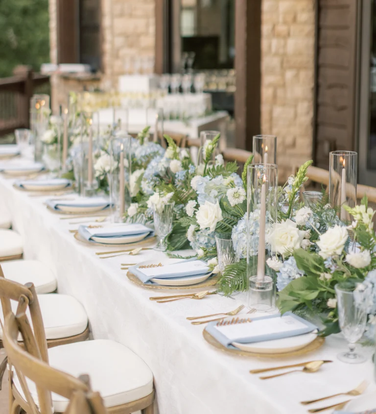 Elegant outdoor wedding table with white and blue floral garland, gold flatware, taper candles, and blue napkins