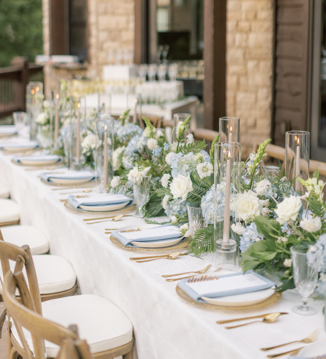 Elegant outdoor wedding table with white and blue floral garland, gold flatware, taper candles, and blue napkins