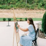Artist in blue dress painting outdoor landscape on easel at wedding venue