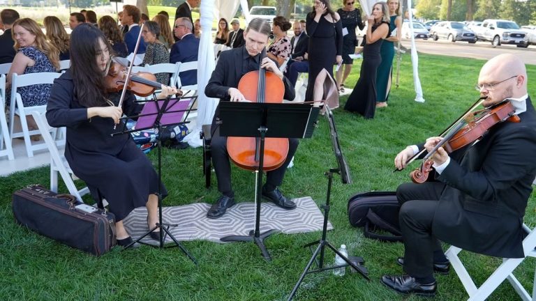 Three-piece string ensemble playing violin and cello for seated wedding ceremony attendees