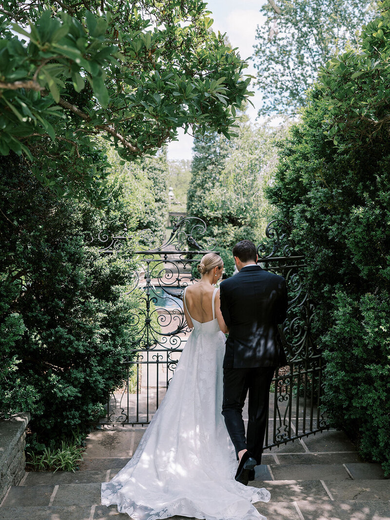 Bride in backless lace gown with groom approaching ornate garden gate beneath canopy of trees