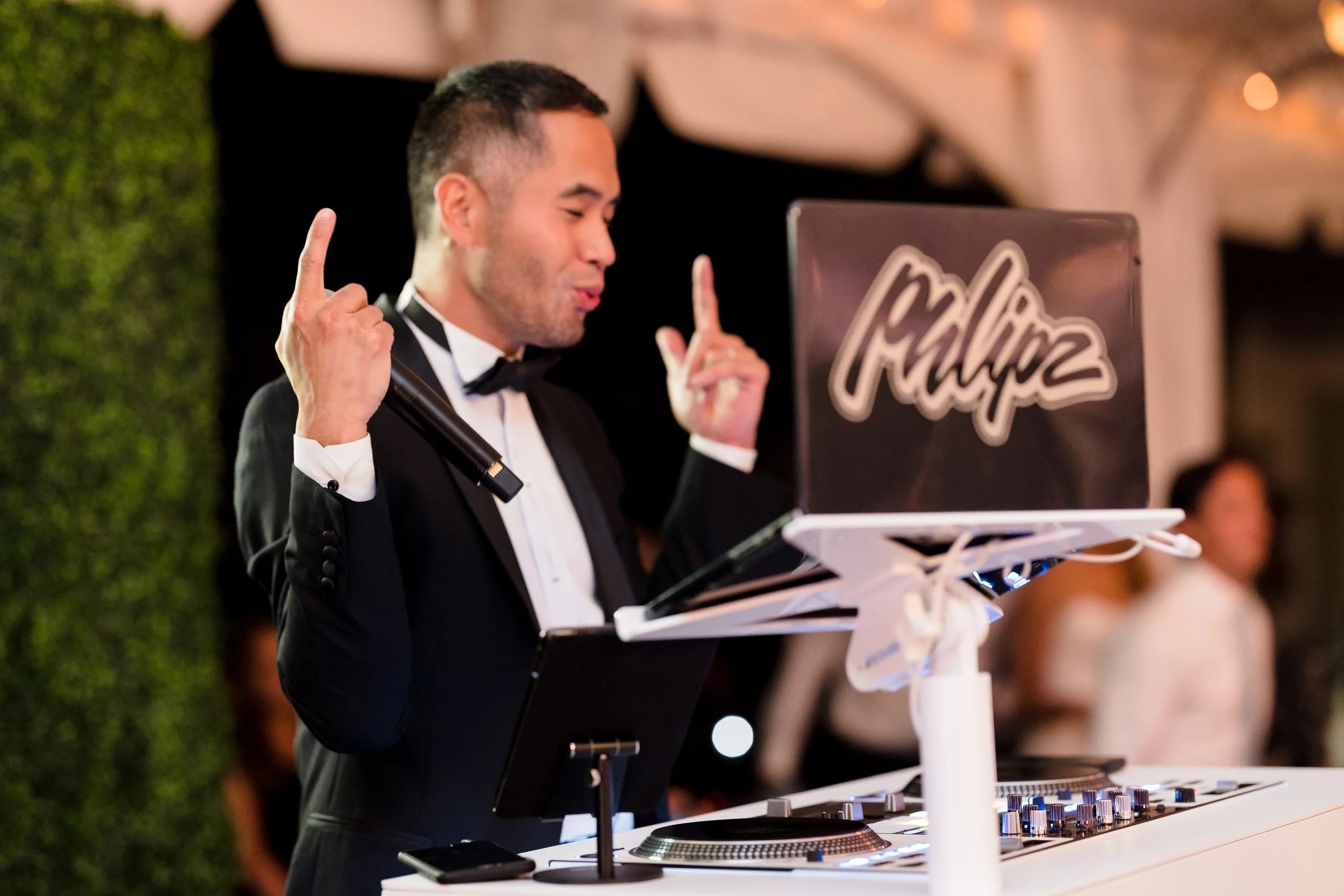 DJ in formal attire gesturing expressively while performing at event with illuminated "Philipz" sign