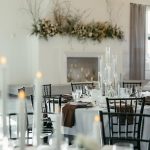 Elegant wedding reception table with black chiavari chairs, tall glass candelabras, and greenery garland above white fireplace mantel
